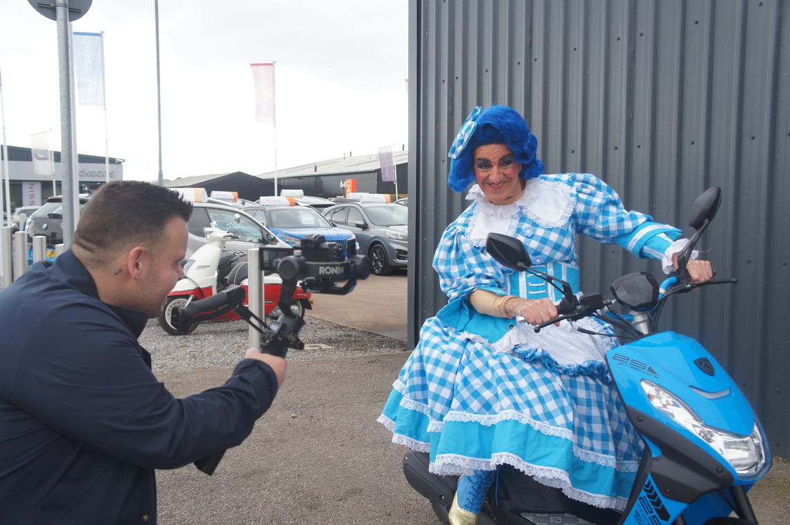 Dame Moira sits on a blue scooter, pretending to ride it, while Dickson's film team record it.